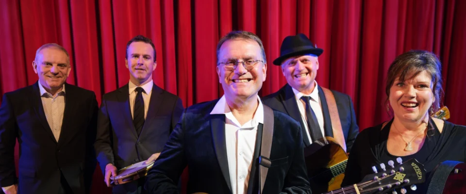 Five band members smiling on stage with red velvet curtains in background.