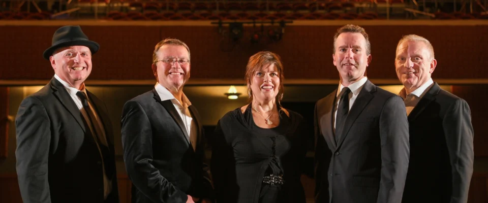 Five band members in a theatre with wood and lights in background.
